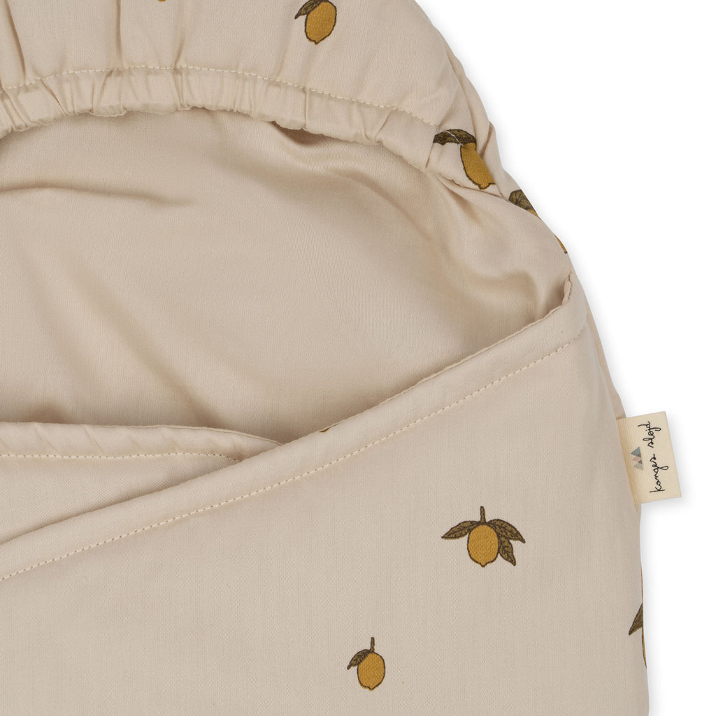 Close-up of the Konges Sloejd Doll Sleeping Bag - Lemon, featuring a beige organic cotton fabric with a lemon print, elastic edge, and a small fabric tag on the right.