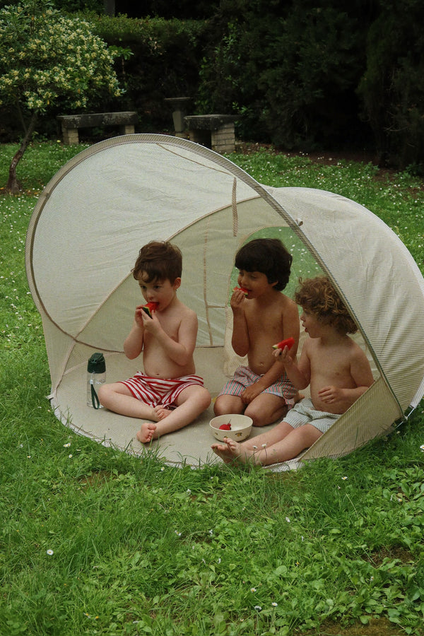 Three young children sit in the Konges Sloejd Pop-Up Beach Tent UPF 50+ on grassy ground, eating watermelon and enjoying a playful, sun-safe summer day outdoors surrounded by greenery.