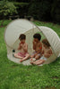 Three young children sit in the Konges Sloejd Pop-Up Beach Tent UPF 50+ on grassy ground, eating watermelon and enjoying a playful, sun-safe summer day outdoors surrounded by greenery.