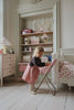 A young girl plays at a small table in a cozy bedroom decorated with shelves, toys, and the Konges Sloejd High Chair - Rosie Rose, featuring scallop edges. The tidy, pastel room is softly lit for a warm and inviting atmosphere.