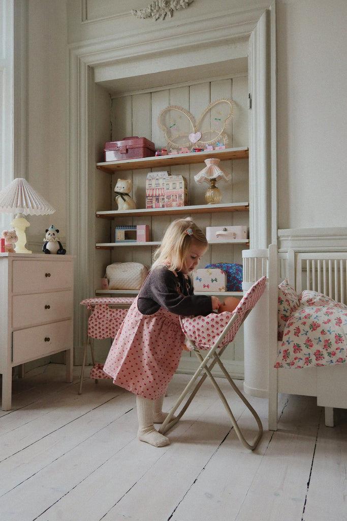 A young girl plays at a small table in a cozy bedroom decorated with shelves, toys, and the Konges Sloejd High Chair - Rosie Rose, featuring scallop edges. The tidy, pastel room is softly lit for a warm and inviting atmosphere.