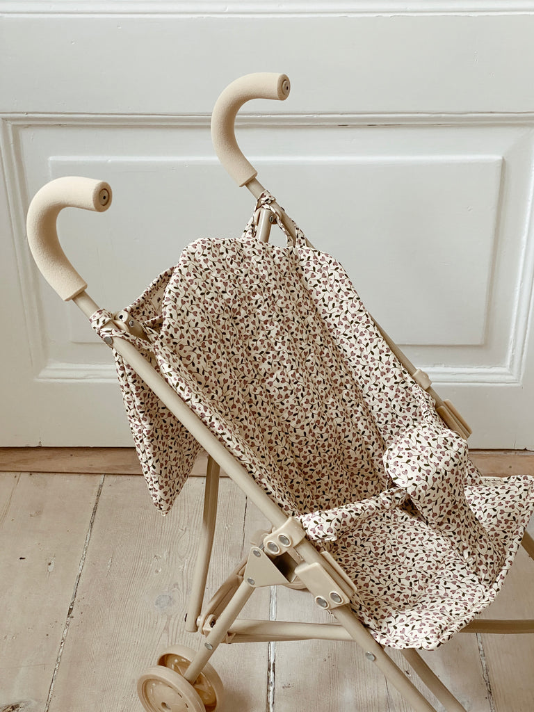 The Konges Sloejd Doll Stroller - Milk Tank, featuring soft handles and a floral-patterned fabric seat in beige, is positioned on a light wooden floor in front of a white paneled door. Its double wheels offer enhanced stability, making it ideal for playtime adventures.