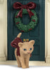A small tan cat with a red ribbon tied around its neck stands in front of a Konges Sloejd Christmas Door, which is a miniature dark green door decorated with a Christmas wreath.