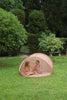 Two young children sit inside a Konges Sloejd Lace Pop-Up Beach Tent UPF 50+ on a grassy lawn, surrounded by lush green trees and bushes, sharing a drink and snacks together.