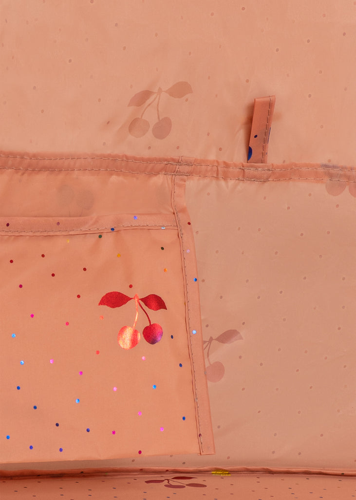 Close-up of the Konges Sloejd Lace Pop-Up Beach Tent UPF 50+ in peach fabric with small polka dots, UV protection, a shiny red cherry graphic, a pocket with multi-colored specks, and a fabric loop above the pocket.