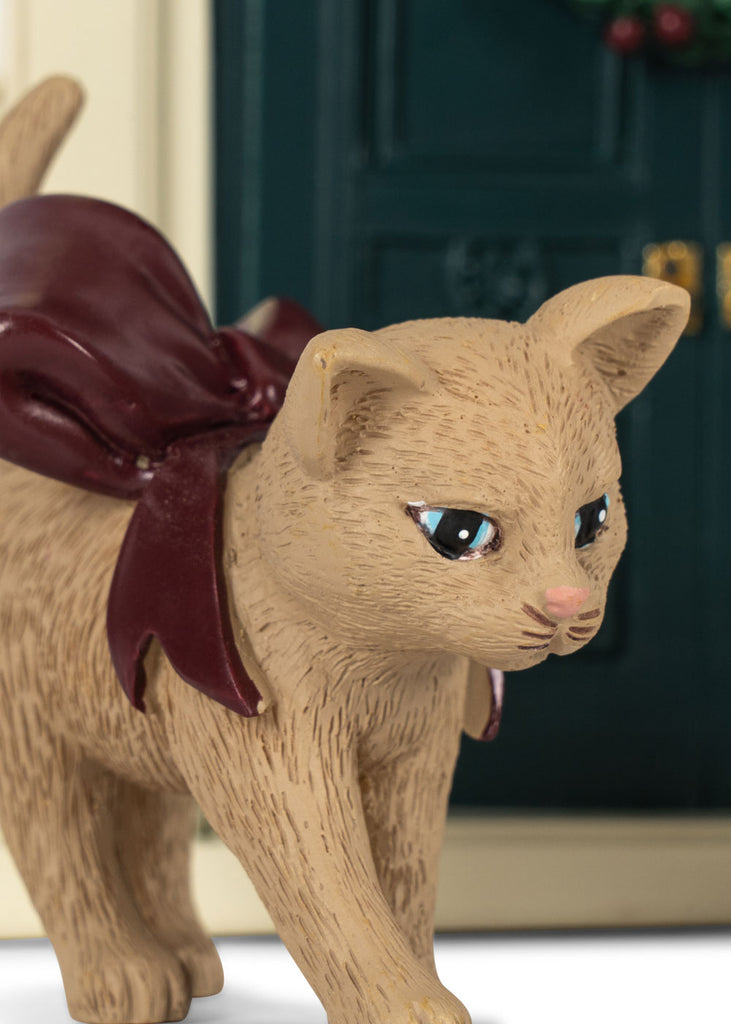 A small, beige toy cat with a large, dark red ribbon on its back, standing in front of a Konges Sloejd Christmas Door.