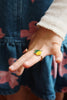 A child in a blue dress with pink flowers and a white long-sleeve shirt shows tiny fingers wearing a colorful ring from the Konges Sloejd 12 Pack Ring Set, featuring playful lemon-shaped designs.