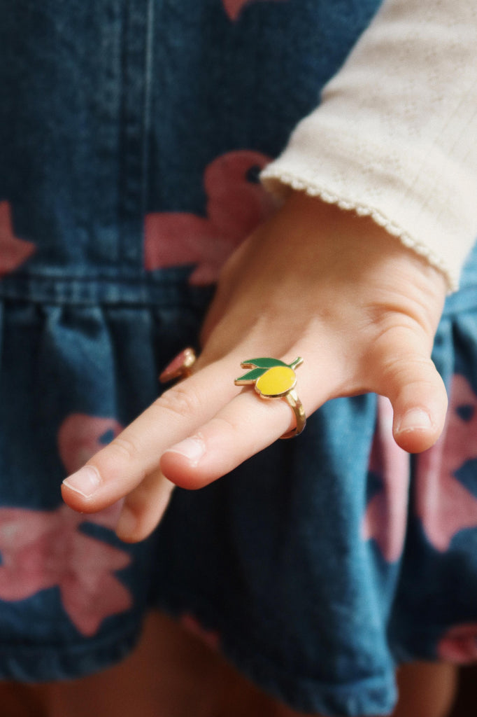 A child in a blue dress with pink flowers and a white long-sleeve shirt shows tiny fingers wearing a colorful ring from the Konges Sloejd 12 Pack Ring Set, featuring playful lemon-shaped designs.