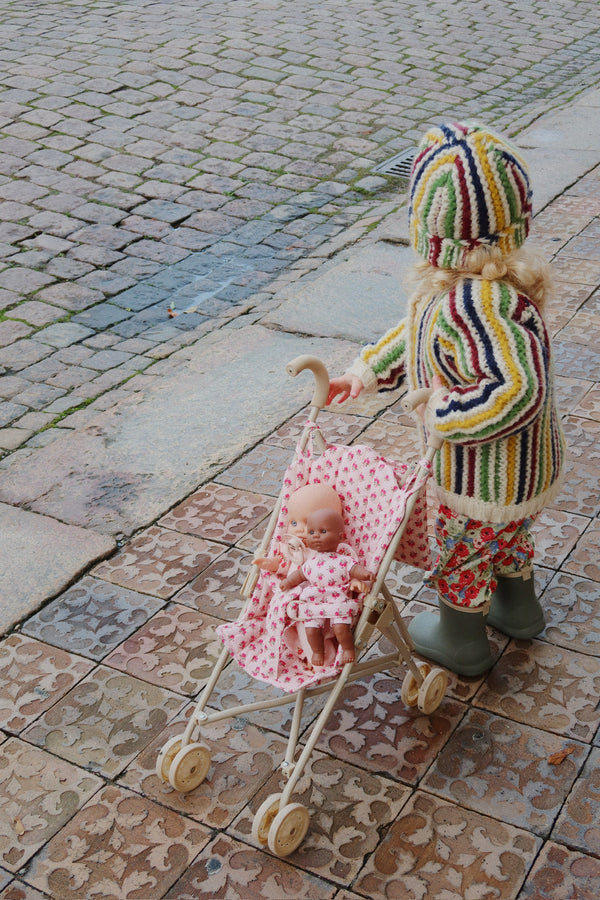 A child in a striped sweater, floral pants, and rain boots pushes the Konges Sloejd Doll Stroller - Rosie Rose filled with adorable doll accessories along a patterned sidewalk beside a cobblestone street.
