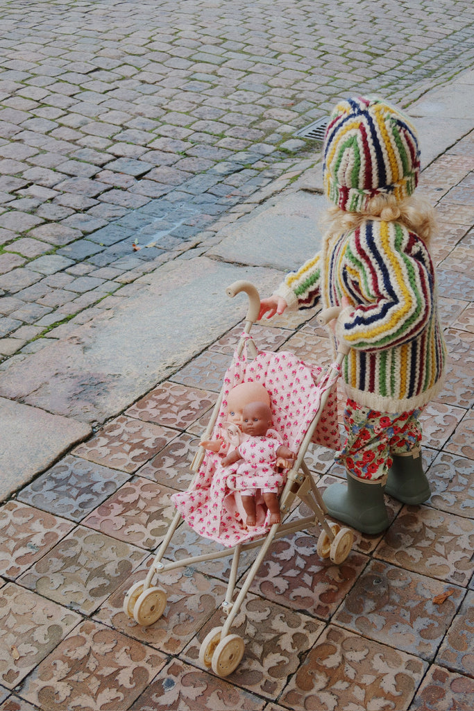 A child in a striped sweater, floral pants, and rain boots pushes the Konges Sloejd Doll Stroller - Rosie Rose filled with adorable doll accessories along a patterned sidewalk beside a cobblestone street.
