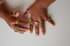 Two small hands with medium brown skin show off the Konges Sloejd 12 Pack Ring Set—multiple colorful, adjustable rings with playful fruit, flower, and heart designs—against a plain white background.