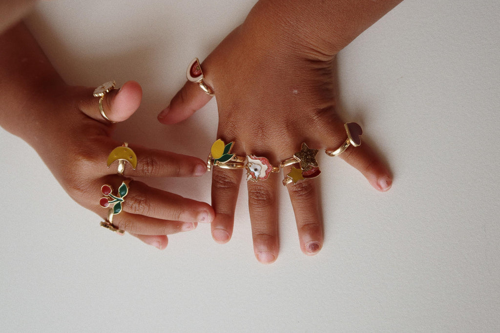 Two small hands with medium brown skin show off the Konges Sloejd 12 Pack Ring Set—multiple colorful, adjustable rings with playful fruit, flower, and heart designs—against a plain white background.