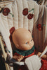 A baby doll with blue eyes sits in the Konges Sloejd Tulle Doll Stroller - Cherry Sequin, featuring red cherry embroidery. The doll wears a red outfit with white polka dots and a green collar.