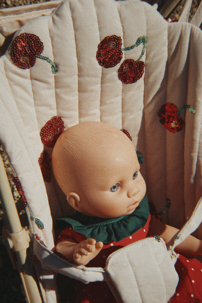 A baby doll with blue eyes sits in the Konges Sloejd Tulle Doll Stroller - Cherry Sequin, featuring red cherry embroidery. The doll wears a red outfit with white polka dots and a green collar.