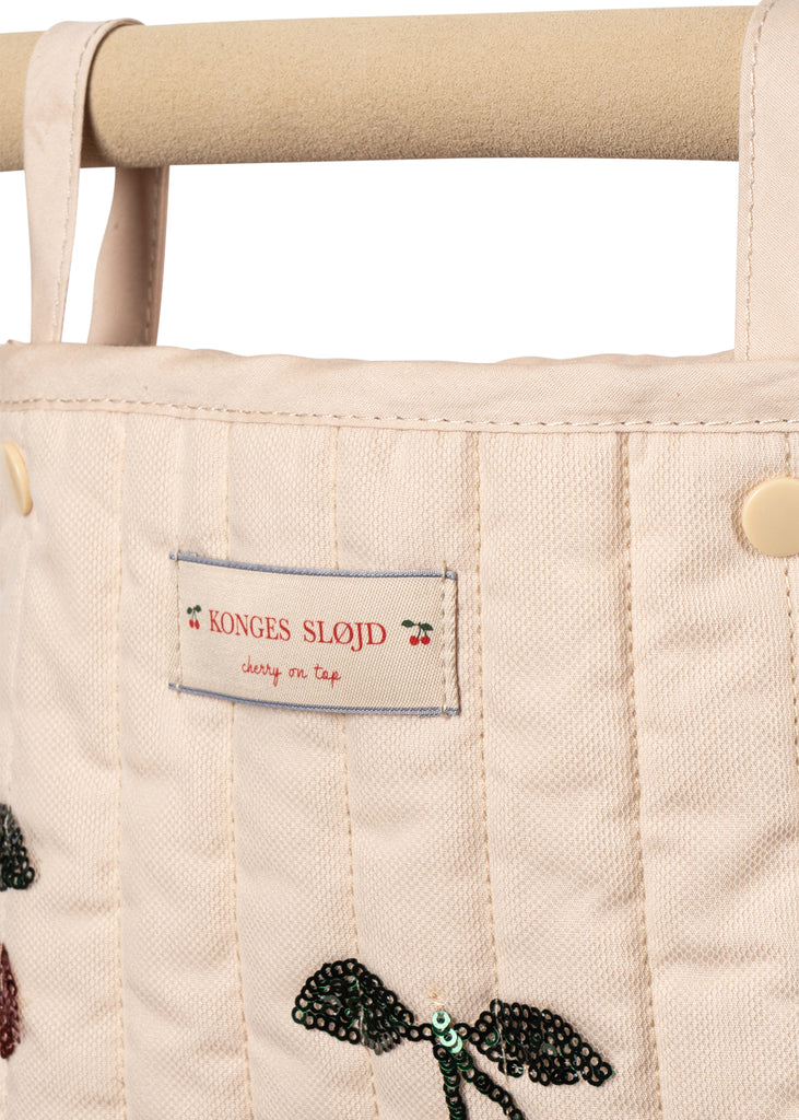 Close-up of the Konges Sloejd Mini Tulle Doll Pram - Cherry Sequin: a cream quilted bag with handles, "Konges Slojd, cherry on top" fabric label, and sequined dragonfly—perfect vintage-style accessory for dolls.
