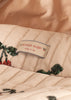 A close-up of quilted fabric on the Konges Sloejd Mini Tulle Doll Pram - Cherry Sequin, showing embroidered green leaves, a white "Konges Slojd cherry on top" label with red cherry graphics, set against a soft beige background.