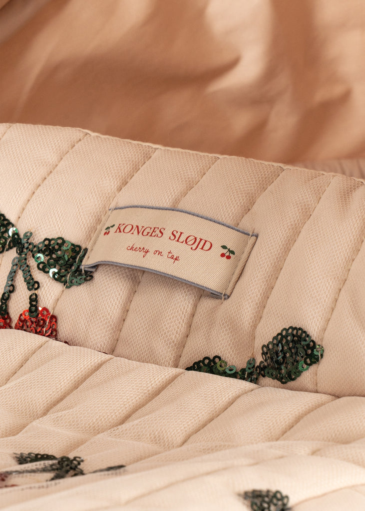 A close-up of quilted fabric on the Konges Sloejd Mini Tulle Doll Pram - Cherry Sequin, showing embroidered green leaves, a white "Konges Slojd cherry on top" label with red cherry graphics, set against a soft beige background.