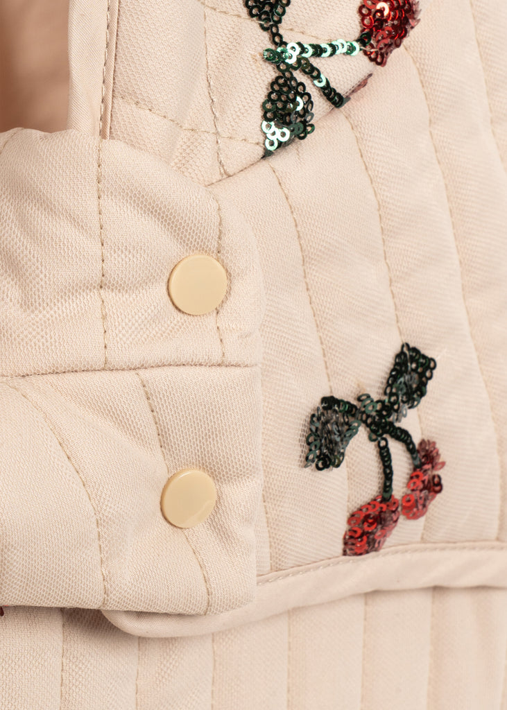 Close-up of the Konges Sloejd Mini Tulle Doll Pram - Cherry Sequin, showing beige quilted fabric with two cream snap buttons and embroidered sequin cherries with green leaves and red fruit.