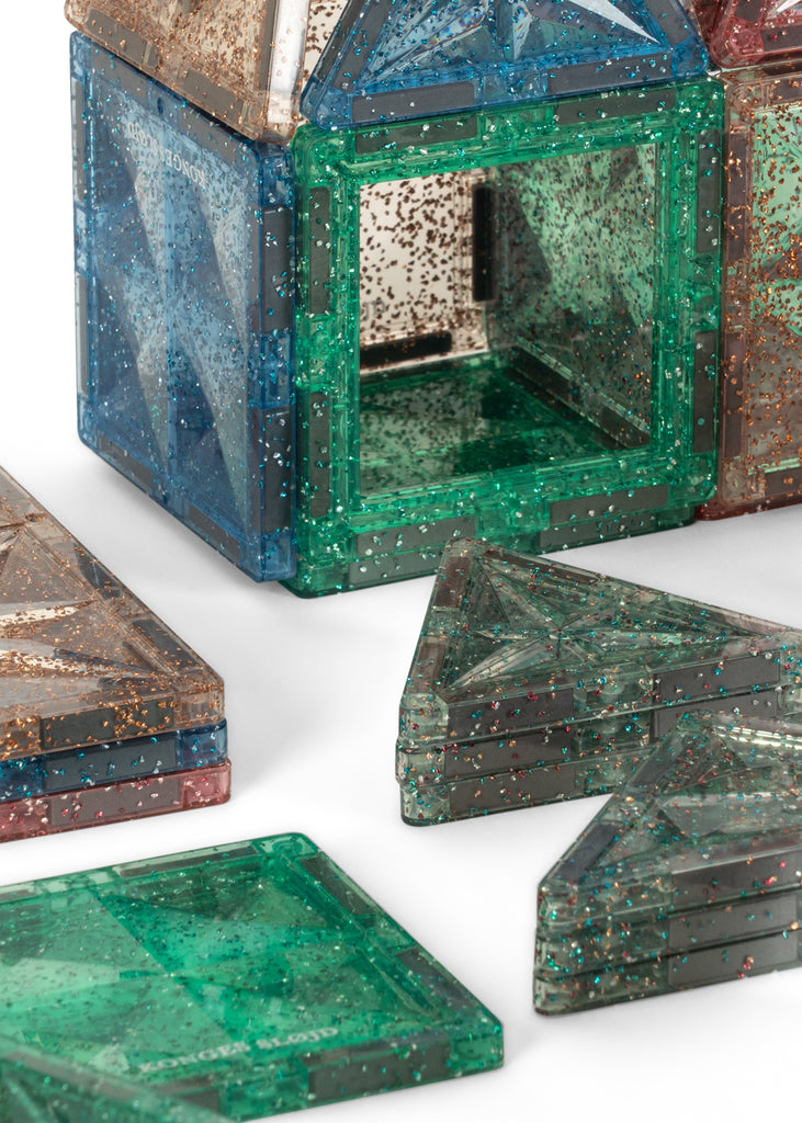 Translucent, glittery magnetic tiles from the Konges Sloejd Piece By Piece Magentic Building Set in square and triangular shapes, some stacked and some arranged into an open cube structure for an educational activity.