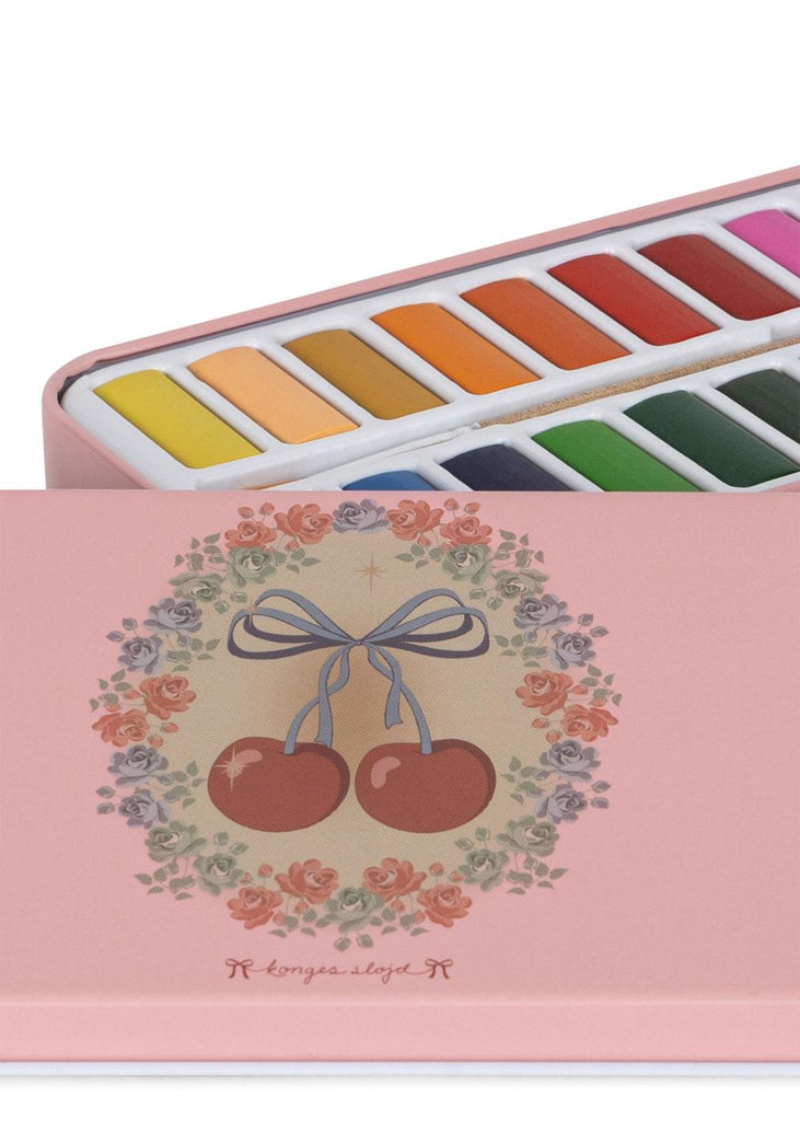The Konges Sloejd Water Colors Paint Set - Cherry Bow comes in a charming pink tin box with a floral and cherry design. The partly open lid reveals neatly arranged colorful paints and a paintbrush inside.