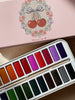 The Konges Sloejd Water Colors Paint Set - Cherry Bow, featuring vibrant color pans and a paint brush, is open on a surface with its pink tin box lid adorned with cherry and floral designs partially visible above the set.