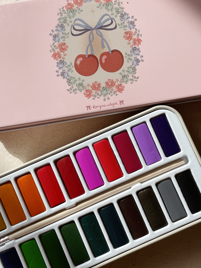 The Konges Sloejd Water Colors Paint Set - Cherry Bow, featuring vibrant color pans and a paint brush, is open on a surface with its pink tin box lid adorned with cherry and floral designs partially visible above the set.