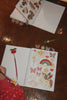 A person writes in a Konges Sloejd My Stationery Box - Unicorn A5 notebook filled with colorful unicorn, rainbow, and star-shaped stickers, with a cherry-topped glitter gel pen nearby.