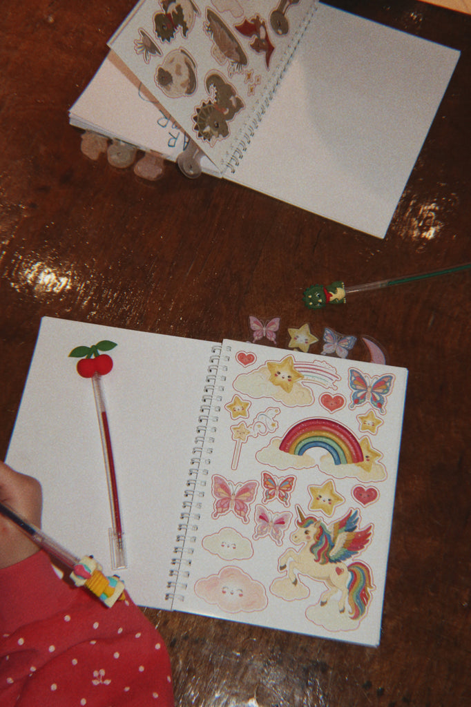A person writes in a Konges Sloejd My Stationery Box - Unicorn A5 notebook filled with colorful unicorn, rainbow, and star-shaped stickers, with a cherry-topped glitter gel pen nearby.