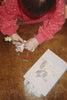 A child in a red polka-dot sweatshirt writes with a glitter gel pen on a unicorn-shaped notepad from the Konges Sloejd My Stationery Box - Unicorn, placed next to an A5 notebook featuring a unicorn illustration.