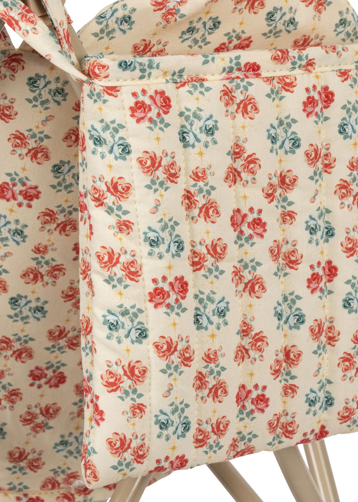 The Konges Sloejd Doll Stroller - Lumina features a padded fabric organizer with a red and teal floral pattern on cream, shown draped over a white wooden chair.