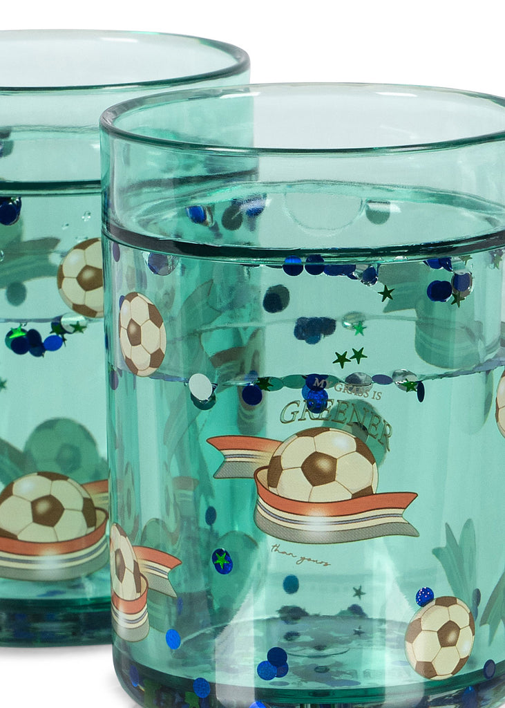 The Konges Sloejd Glitter Cup - Kick It features green-tinted glass, floating soccer balls, stars, and confetti-like blue and green dots—making it a fun and playful addition to any child’s drinkware collection.