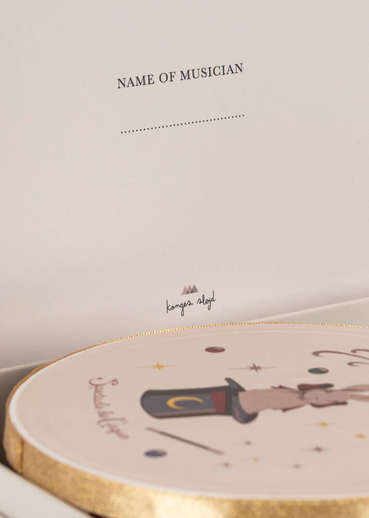 A close-up of a card with "Name of Musician" and a dotted line, shown above an illustrated round box adorned with a magic hat, wand, and stars from the Konges Sloejd Music Set - Circus—perfect for labeling your wooden instruments or adding to your music instrument collection.