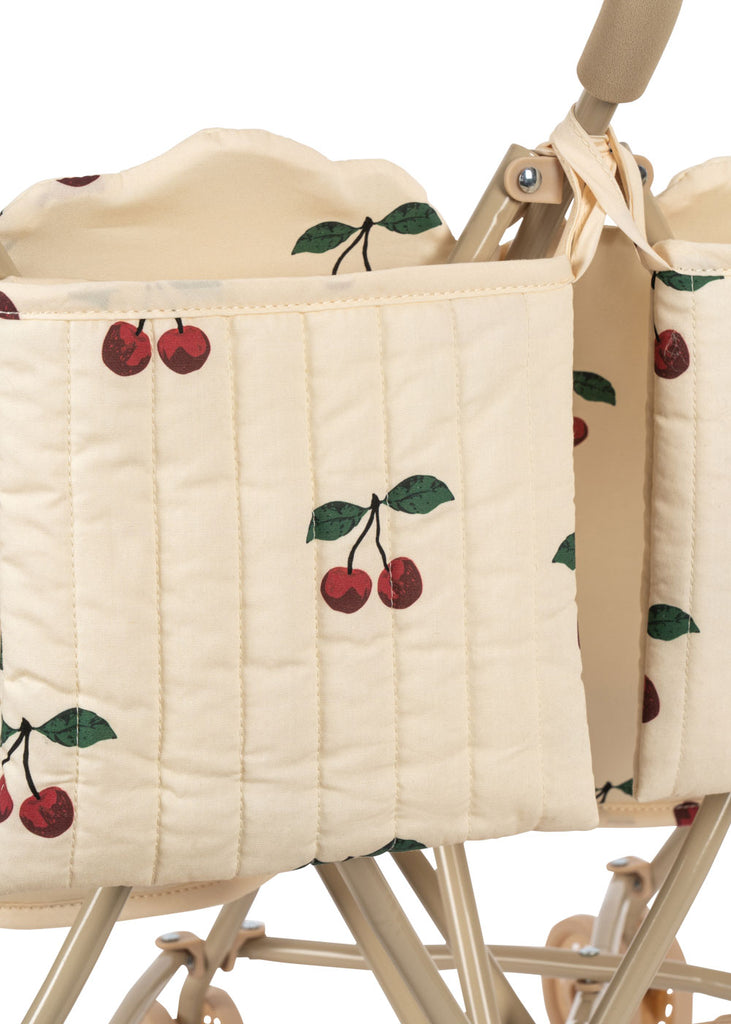 Close-up of the Konges Sloejd Twin Doll Stroller - Cherry, featuring beige quilted fabric with red cherry and green leaf prints, plus tan handles and wheels visible at the bottom.