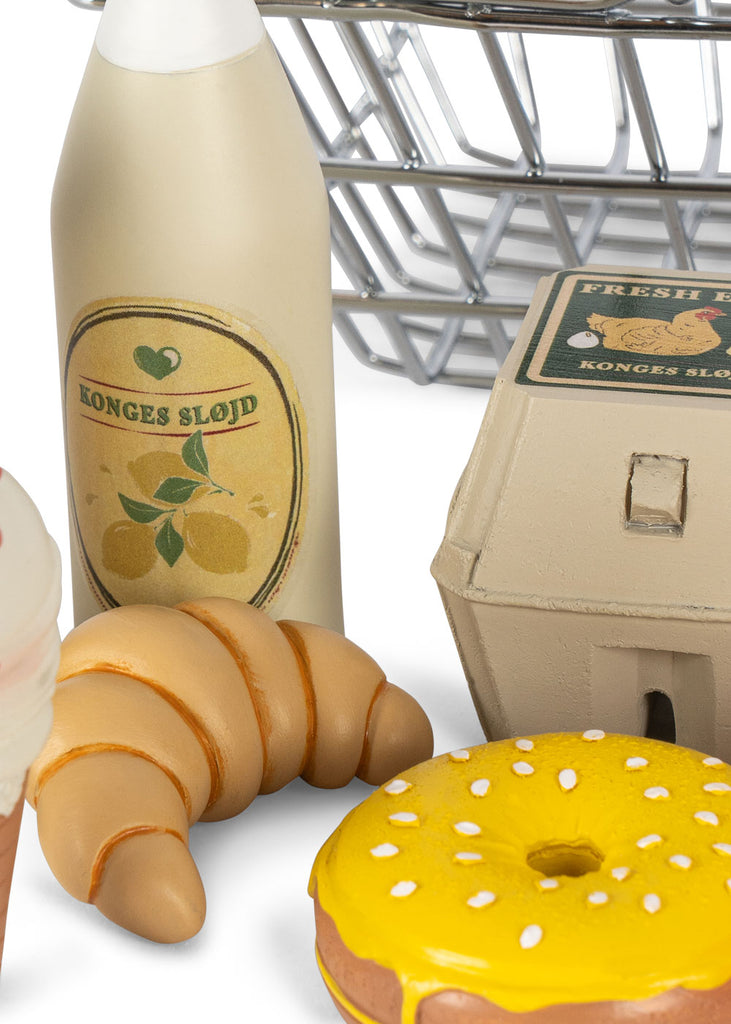 The Konges Sløjd Mini Market Grocery Set includes a sturdy metal basket filled with delightful play food such as a croissant, a yellow-sprinkled donut, an ice cream cone, a bottle with a lemon label, and an egg carton.