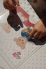 A child’s hands place a teddy bear sticker from the Konges Sloejd My Sticker World Book Set - Teddy onto a polka-dotted page, surrounded by other bear illustrations and floral decorations. Sunlight casts gentle shadows across the paper and the child’s fingers, infusing warmth into this creative sticker activity.