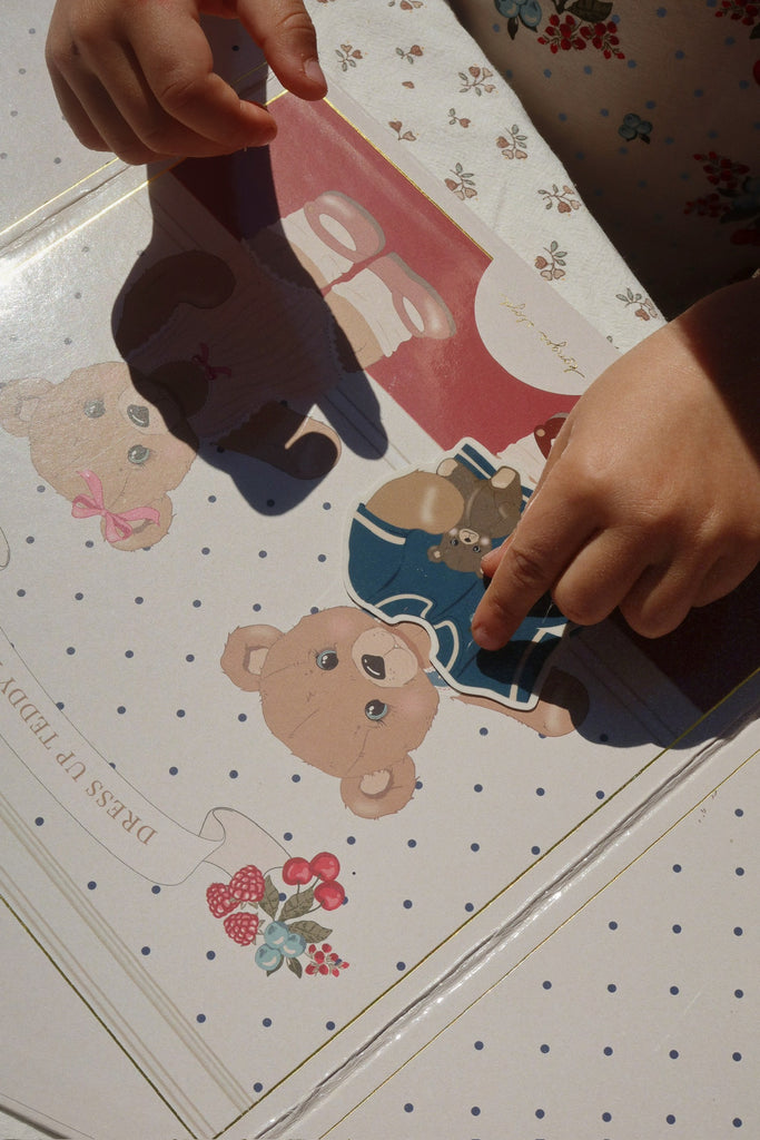 A child’s hands place a teddy bear sticker from the Konges Sloejd My Sticker World Book Set - Teddy onto a polka-dotted page, surrounded by other bear illustrations and floral decorations. Sunlight casts gentle shadows across the paper and the child’s fingers, infusing warmth into this creative sticker activity.