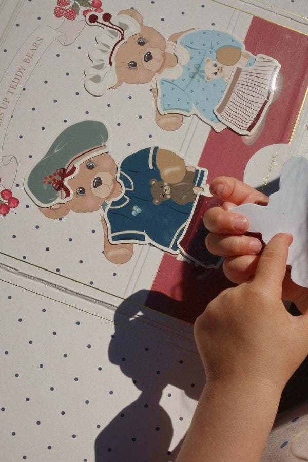 A child’s hands hold a white cutout above a page from the Konges Sloejd My Sticker World Book Set - Teddy, featuring two teddy bear illustrations dressed in clothes against a polka dot background.