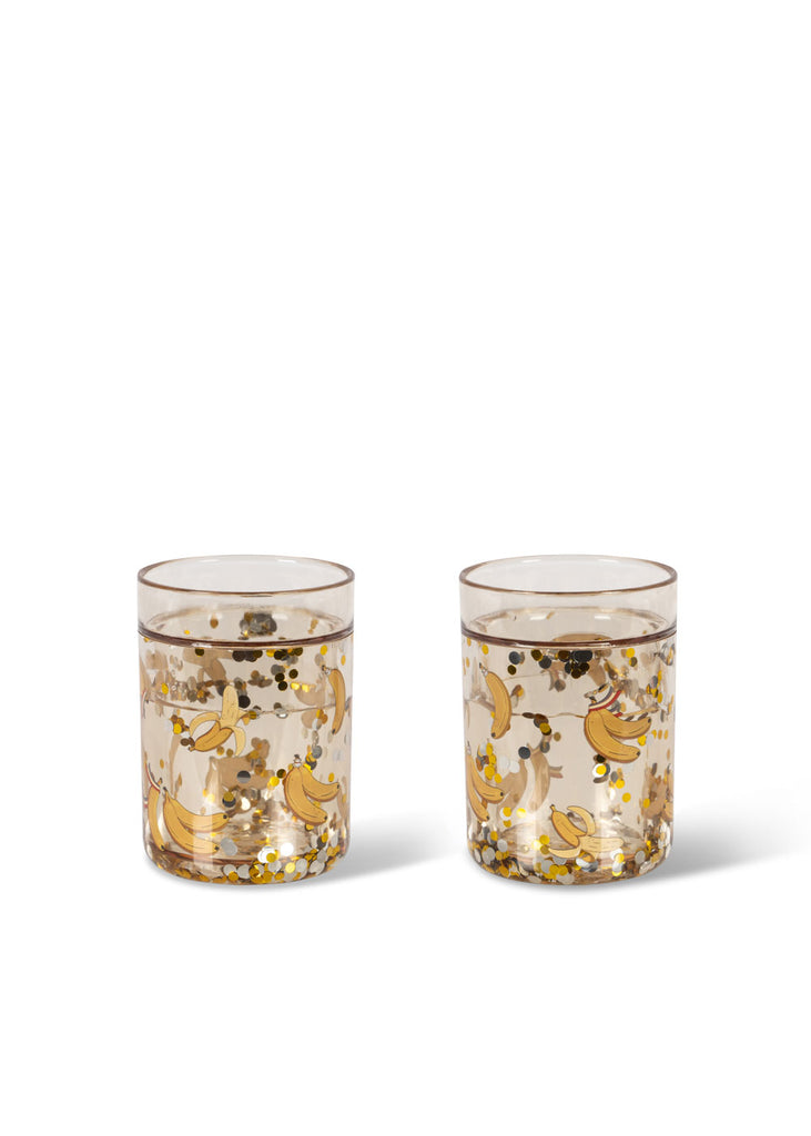 The Konges Sloejd Glitter Cup - Let's Go Bananas features gold and yellow banana and confetti designs, filled with a light-colored drink—an eye-catching choice for playful kids' dinnerware.