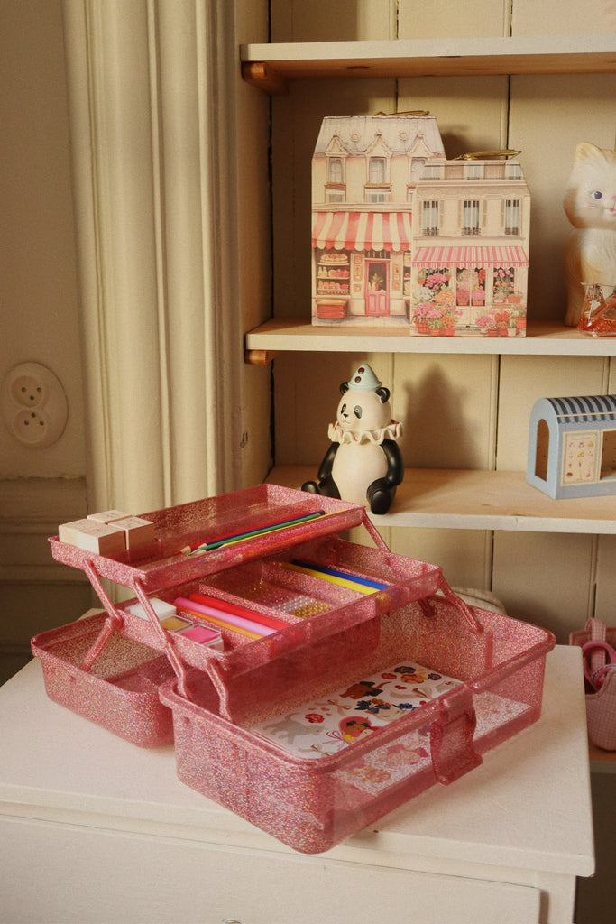 A Konges Sloejd Creative Art Caddy in Glitter Rose sits open on a small table, filled with colored pencils and stickers. Behind it, shelves display a panda figurine, paper house models, and a ceramic cat.