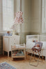 A nursery with light green walls, a crib with stuffed animals, a white dresser with a lamp and teddy bear, the Konges Sloejd High Chair - Kyoto holding a doll, a toy cradle, and a pink dress hanging by the window.