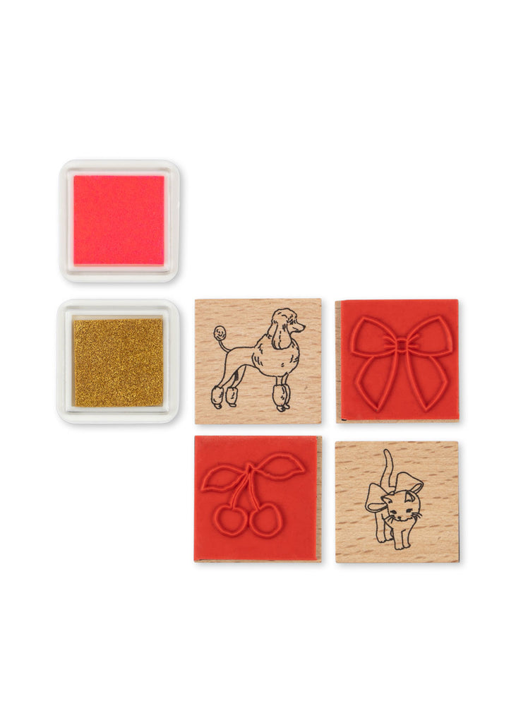 The Konges Sloejd Creative Art Caddy - Glitter Rose includes four charming rubber stamps (poodle, bow, cherries, and cat), along with a pink and a gold ink pad, all beautifully arranged on a white background—making it the perfect addition to any kids' art kit or creative toolbox.