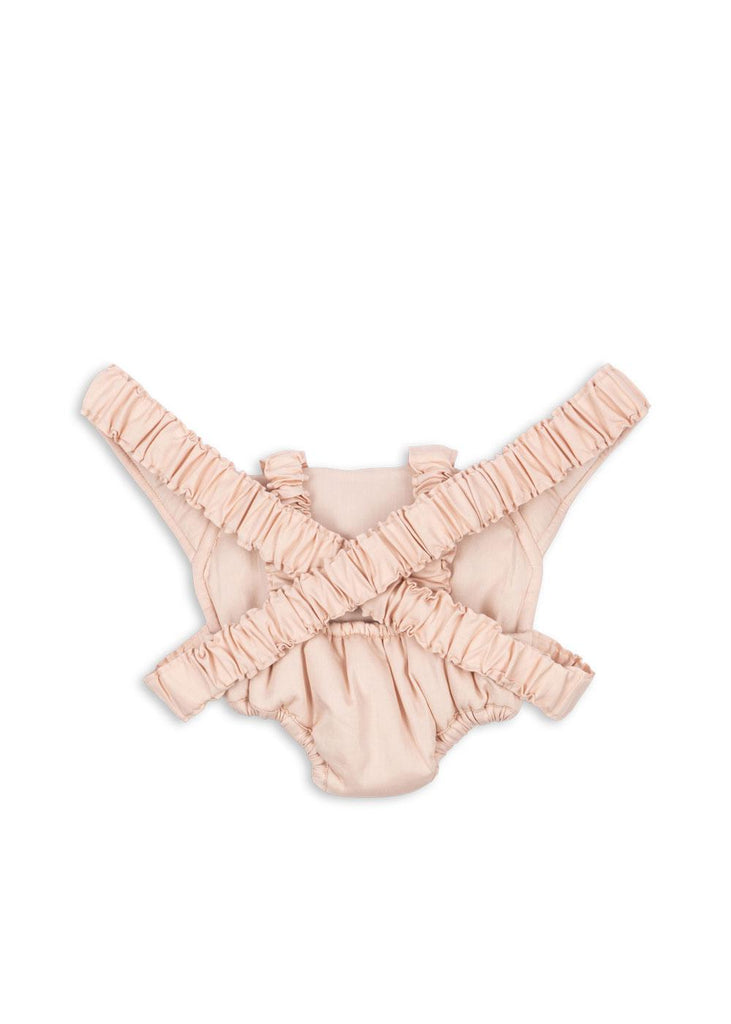 A light pink ruffled baby romper with crisscross straps and elastic leg openings is laid flat beside the Konges Sloejd Doll Carrier - Cameo Rose, creating a sweet matching set for playful moments.