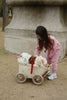 A young child in pink sweats bends over a Konges Sloejd Stoller Kids Wagon - Cherry, filled with stuffed animals— including a white dog, a brown dog, and a pink rabbit— outside on a dirt path.
