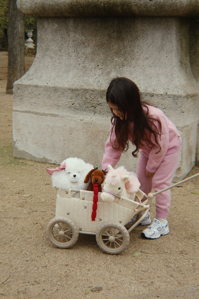 A young child in pink sweats bends over a Konges Sloejd Stoller Kids Wagon - Cherry, filled with stuffed animals— including a white dog, a brown dog, and a pink rabbit— outside on a dirt path.