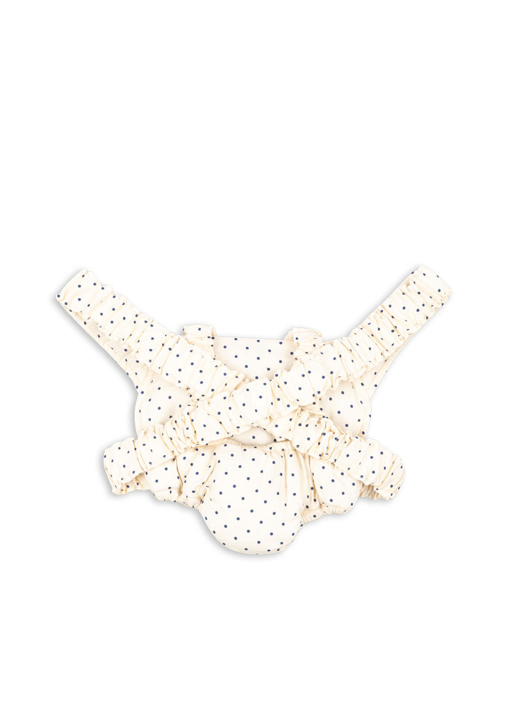 A cream-colored baby diaper cover with navy blue dots, ruffles, and adjustable straps is shown next to the Konges Sloejd Doll Carrier - Estate Dot on a white background.