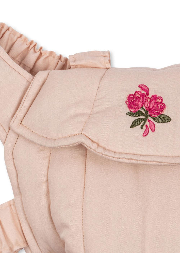 Close-up of the Konges Sloejd Doll Carrier in Cameo Rose, made from light pink cotton, with a ruffled strap and a flap embroidered with two pink flowers and green leaves.