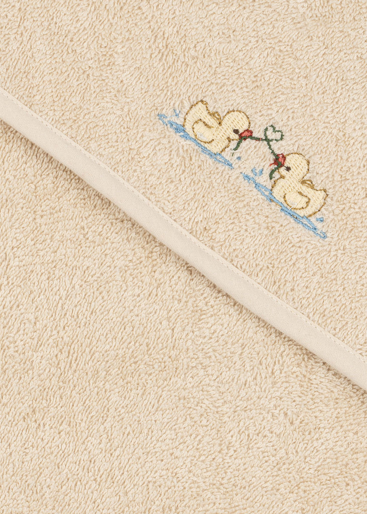 Konges Sloejd Organic Cotton Baby Towel in beige, featuring an embroidered design of two yellow ducks facing each other and holding a green stem with red flowers, displayed on a textured surface with a diagonal seam.