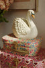The Konges Sloejd Money Bank - Swan, a white ceramic swan, sits on vintage floral and pink heart suitcases—perfect as a charming decoration and for saving coins. Flowers and a framed picture appear in the background.