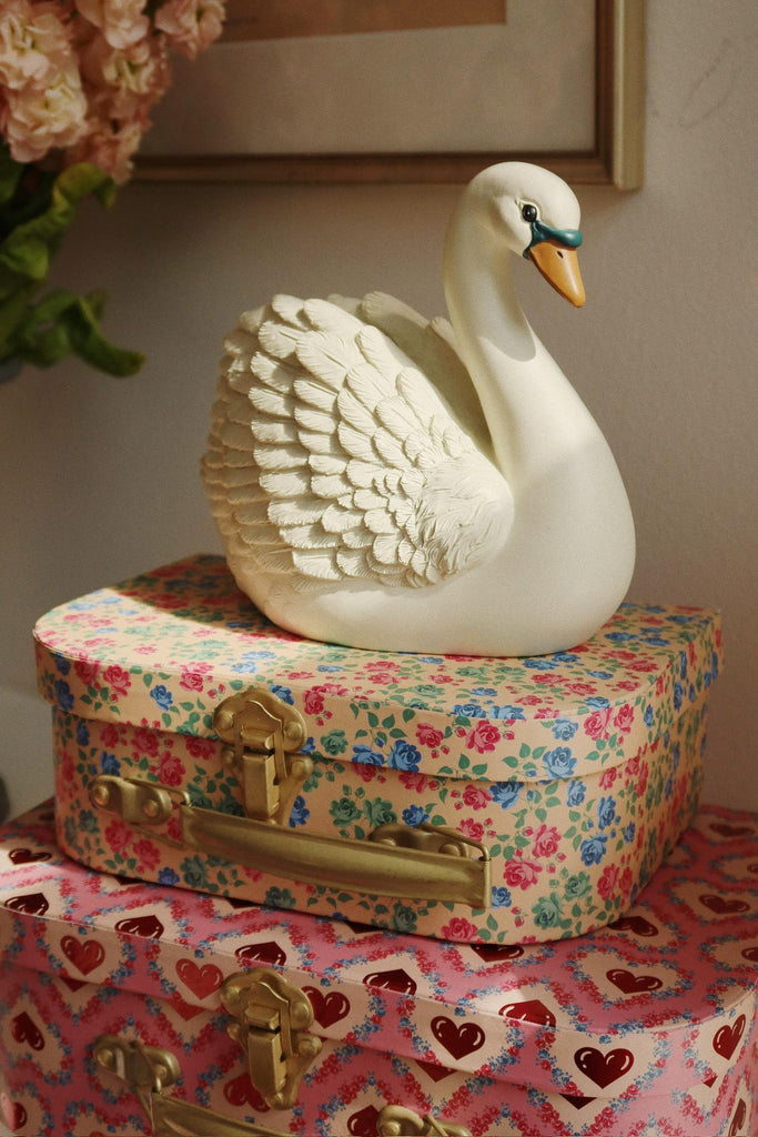The Konges Sloejd Money Bank - Swan, a white ceramic swan, sits on vintage floral and pink heart suitcases—perfect as a charming decoration and for saving coins. Flowers and a framed picture appear in the background.