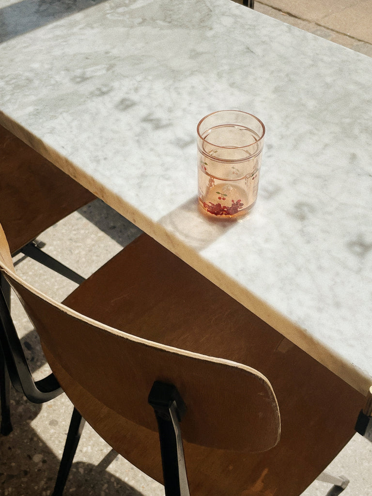 A Konges Sloejd Glitter Cup - Cherry sparkles with a light drink and red berries on a white marble table by a wooden chair, as sunlight highlights the playful charm of these kids cups perfect for any setting.